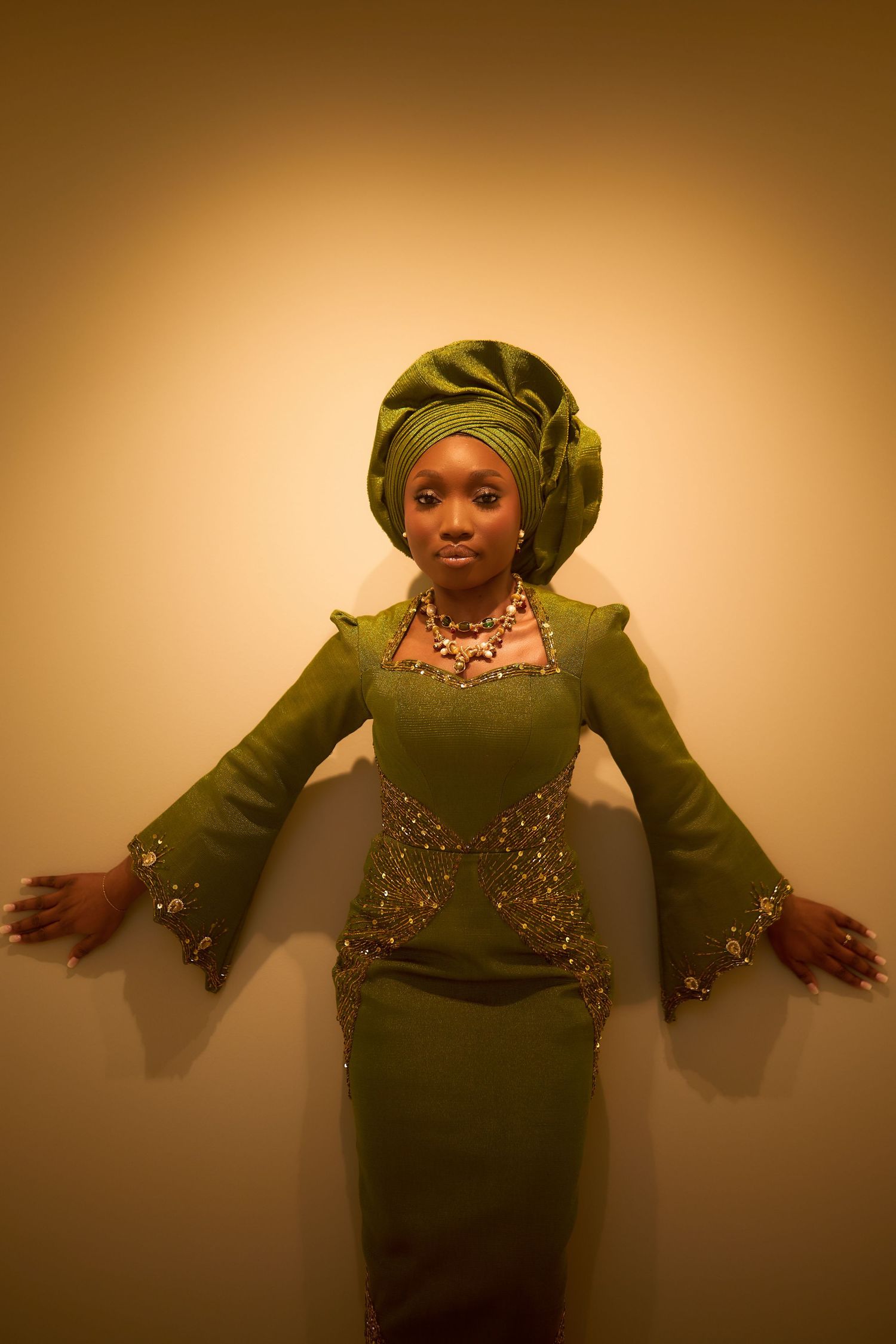 Woman in elegant green Nigerian traditional attire with gele headwrap and gold jewelry posing against warm background.