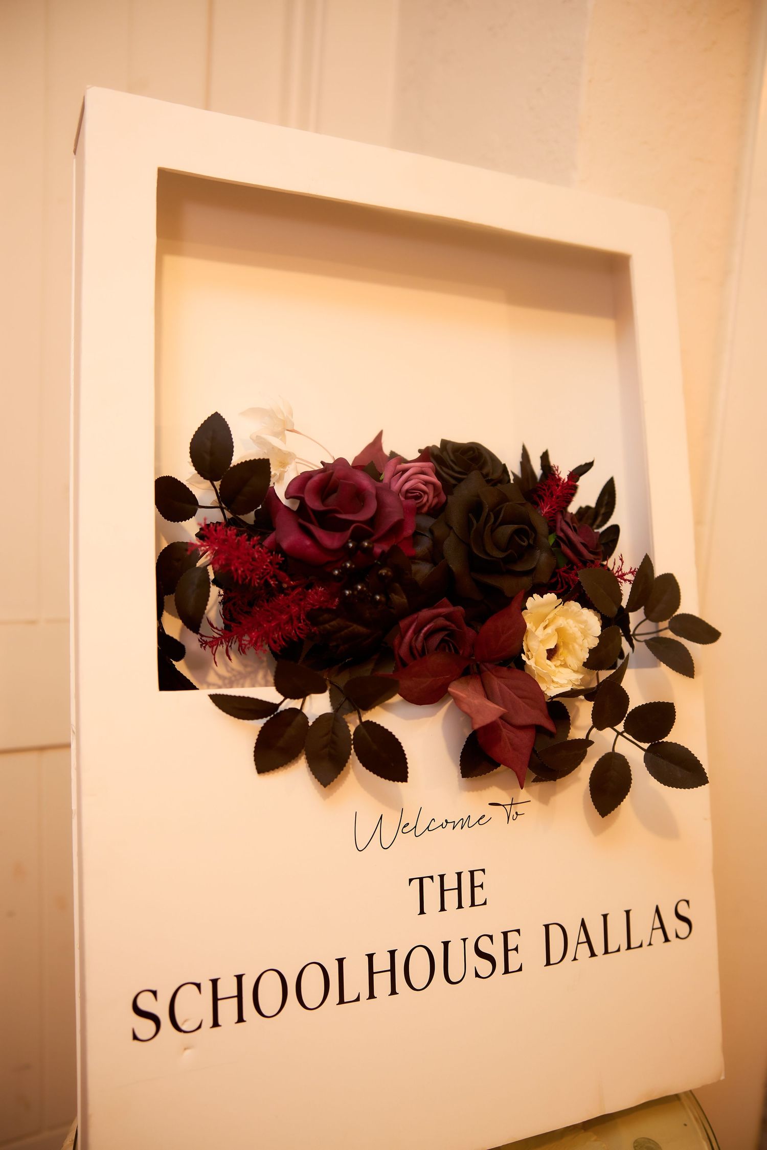 Welcome to The Schoolhouse Dallas sign decorated with dark burgundy and black floral arrangement.