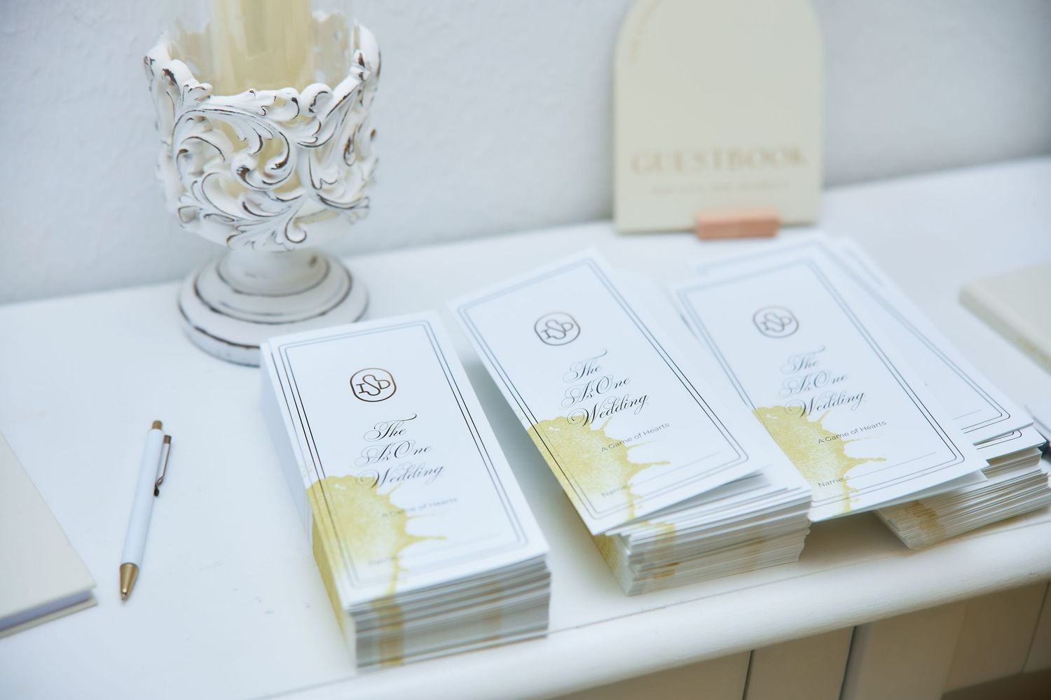 Elegant wedding programs stacked on a table with gold accents next to a decorative white candle holder and pen.