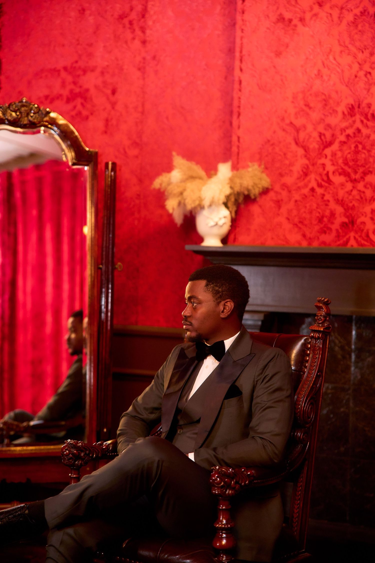 Elegant Black man in tuxedo seated in ornate chair against red damask wall beside gold-framed mirror.
