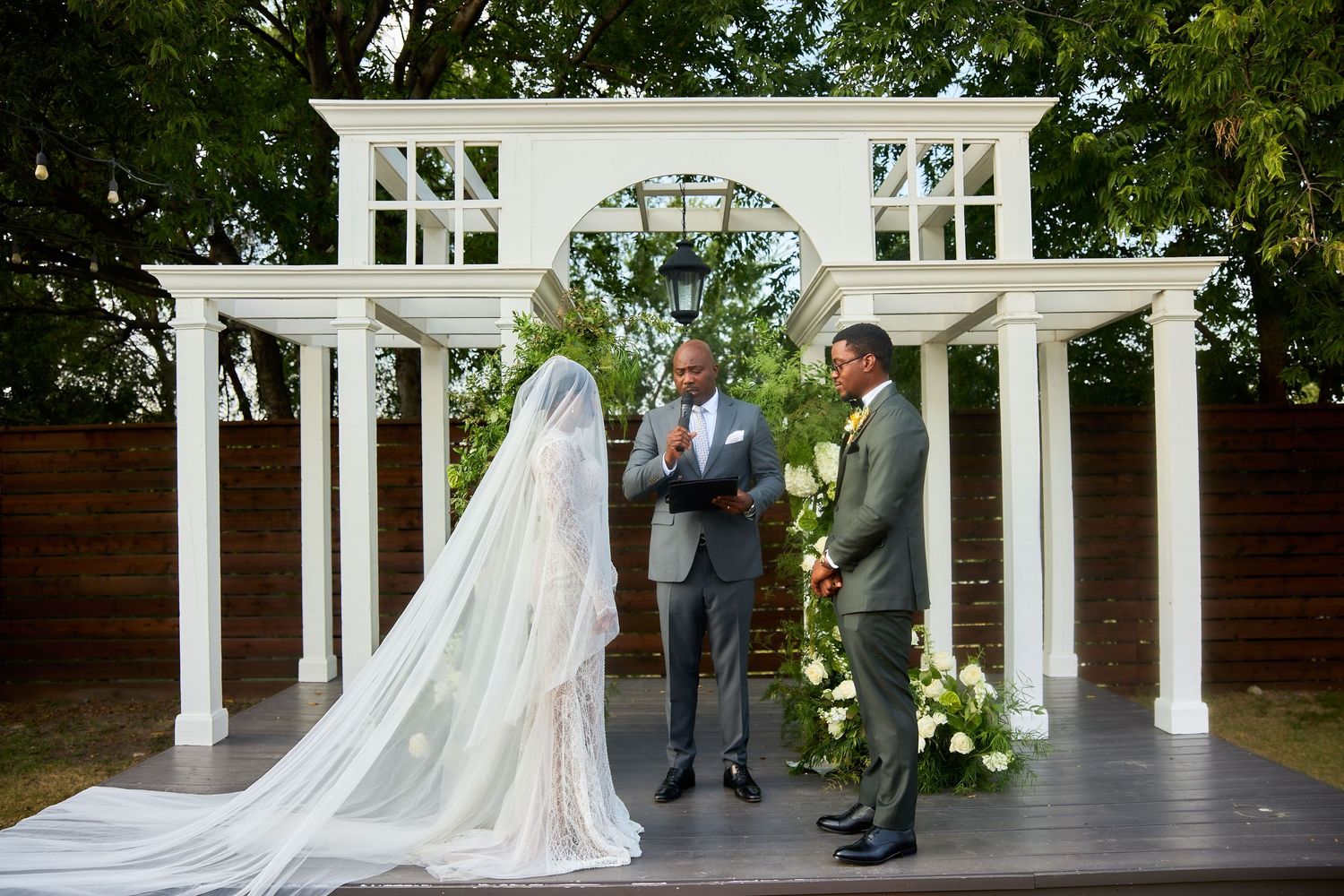 Outdoor wedding ceremony with bride in long veil, groom, and officiant standing under white pergola with floral arrangements.