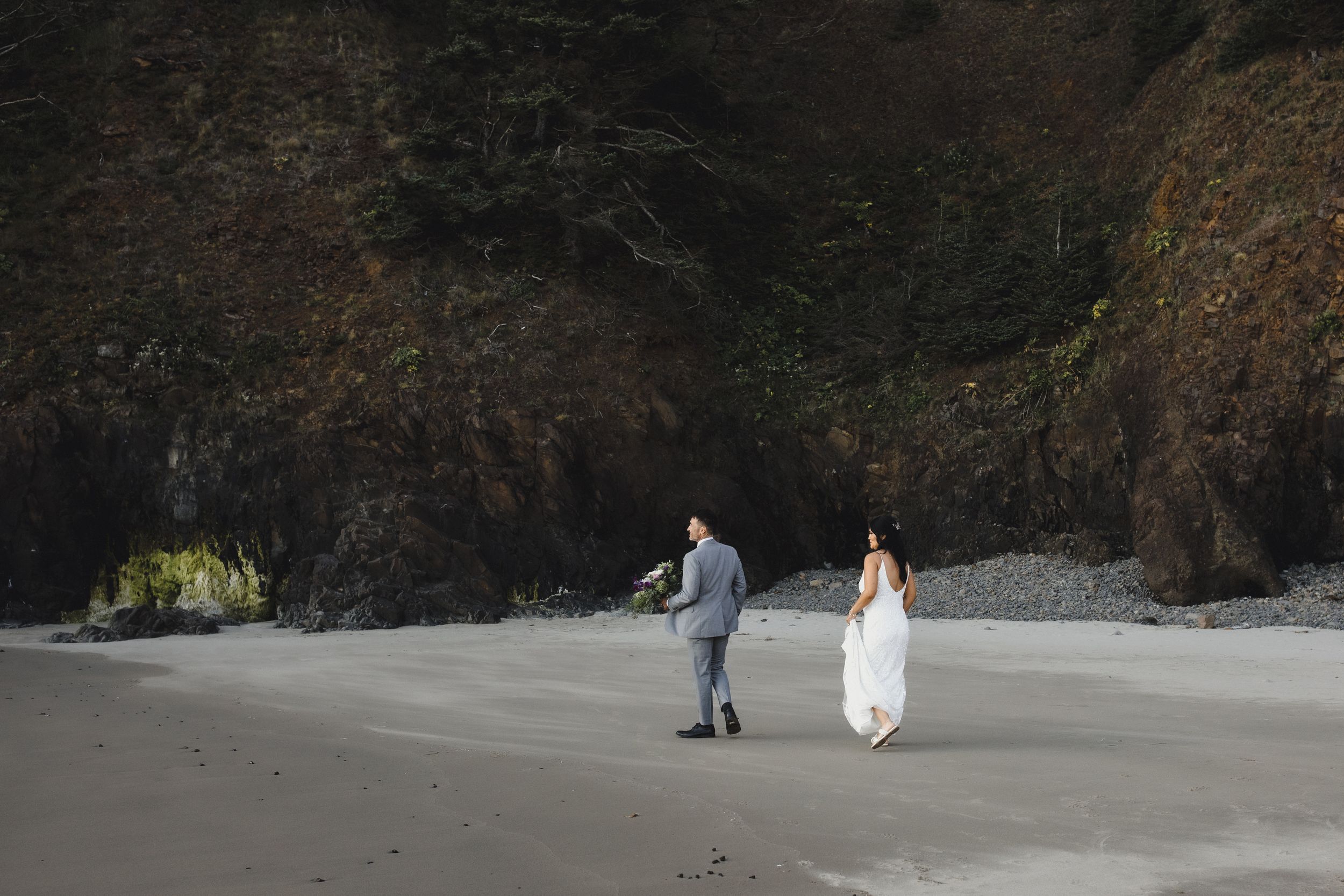 Bride and groom walking hand in hand on a sandy beach with dramatic rocky cliffs in the background.