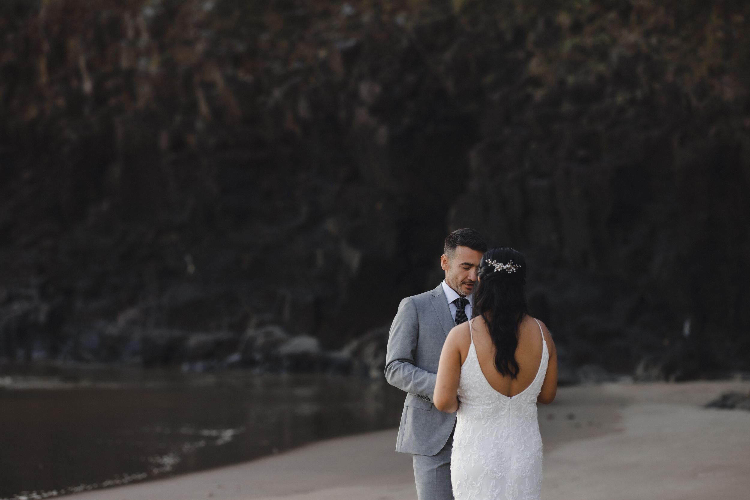 Bride and groom share an intimate moment on a sandy beach with dramatic dark rocky cliffs in the background.