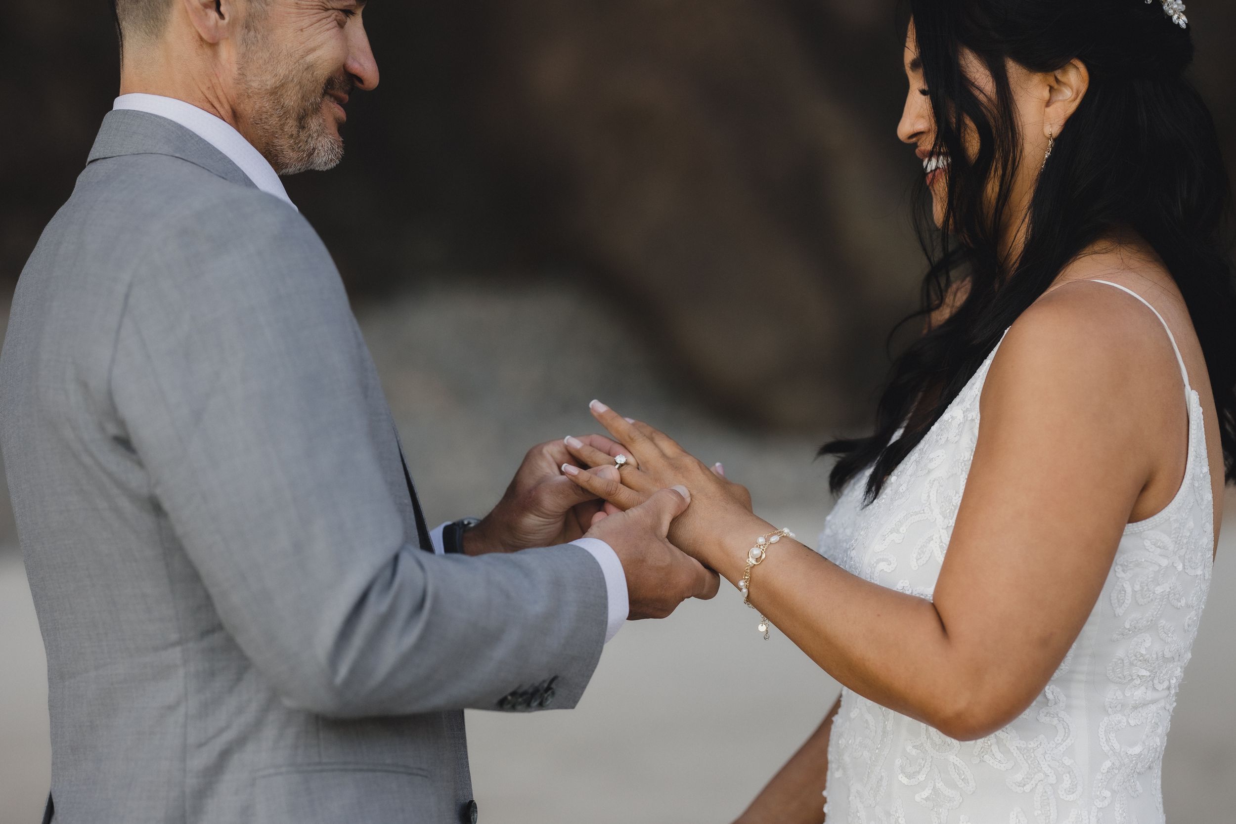 Groom placing wedding ring on bride's finger during outdoor beach ceremony, both smiling joyfully.