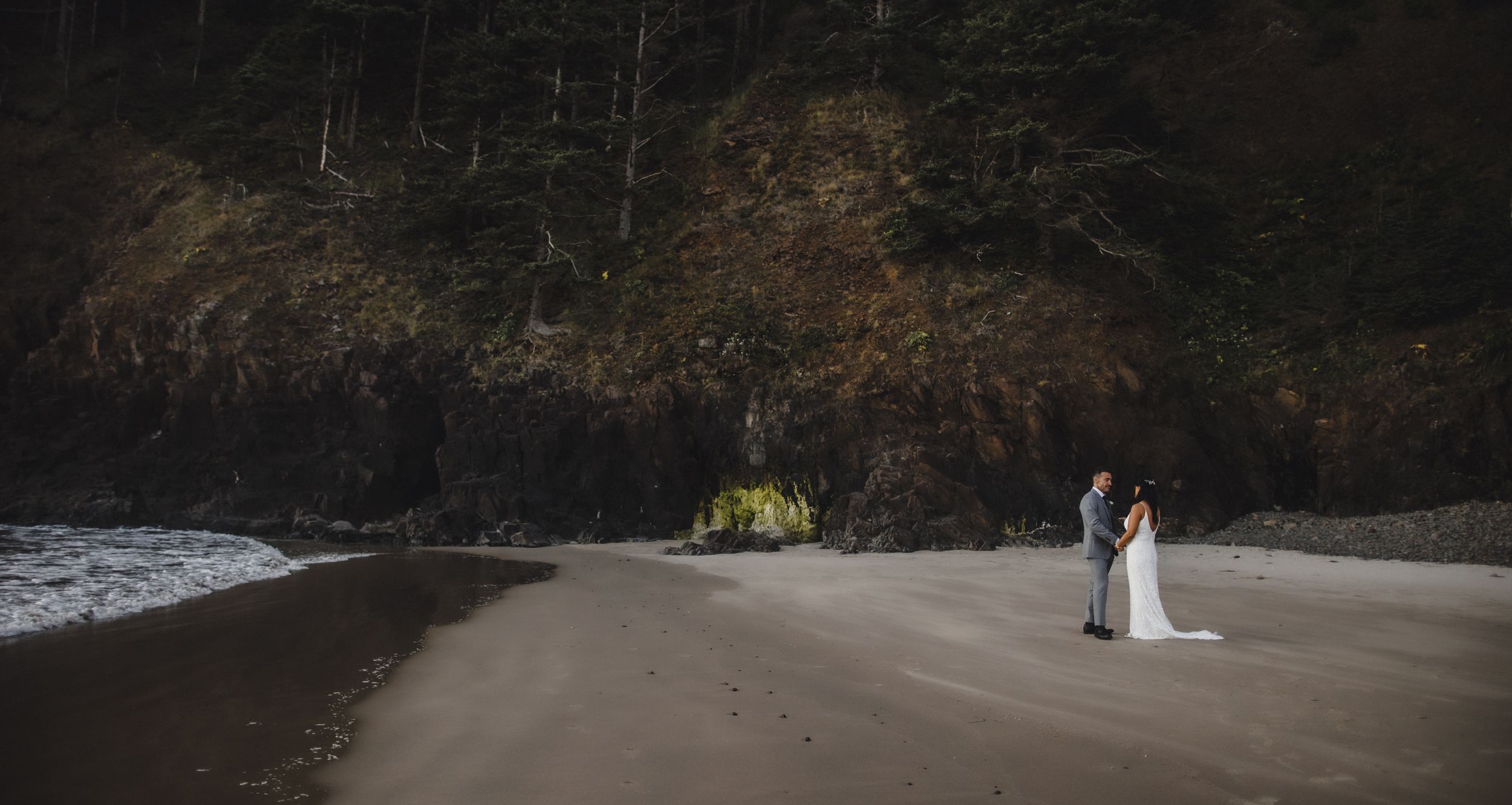 Bride and groom stand together on a dramatic dark sandy beach with towering forested cliffs in the background.