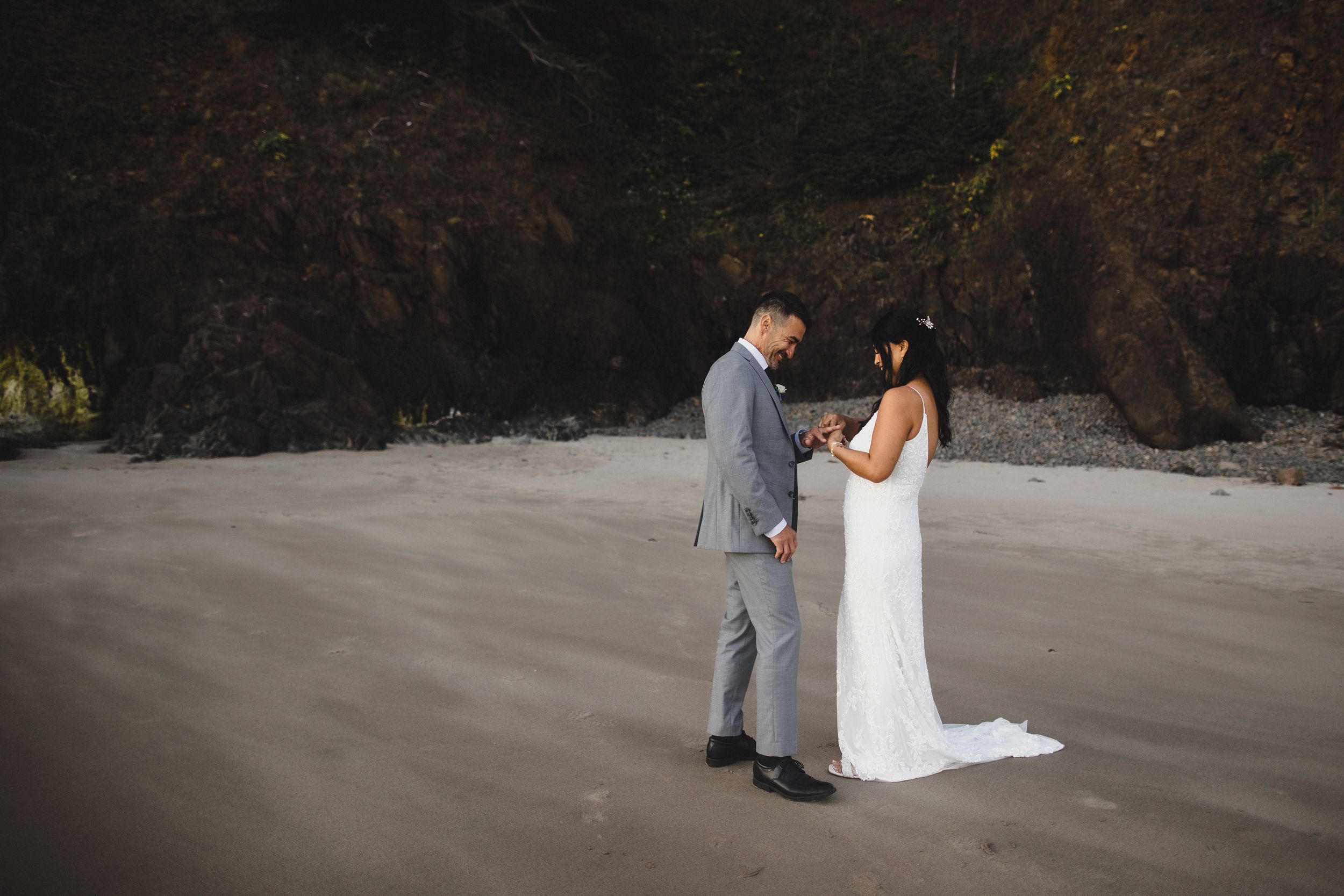 Bride and groom share a romantic moment on a sandy beach, surrounded by dramatic dark cliffs during their wedding.