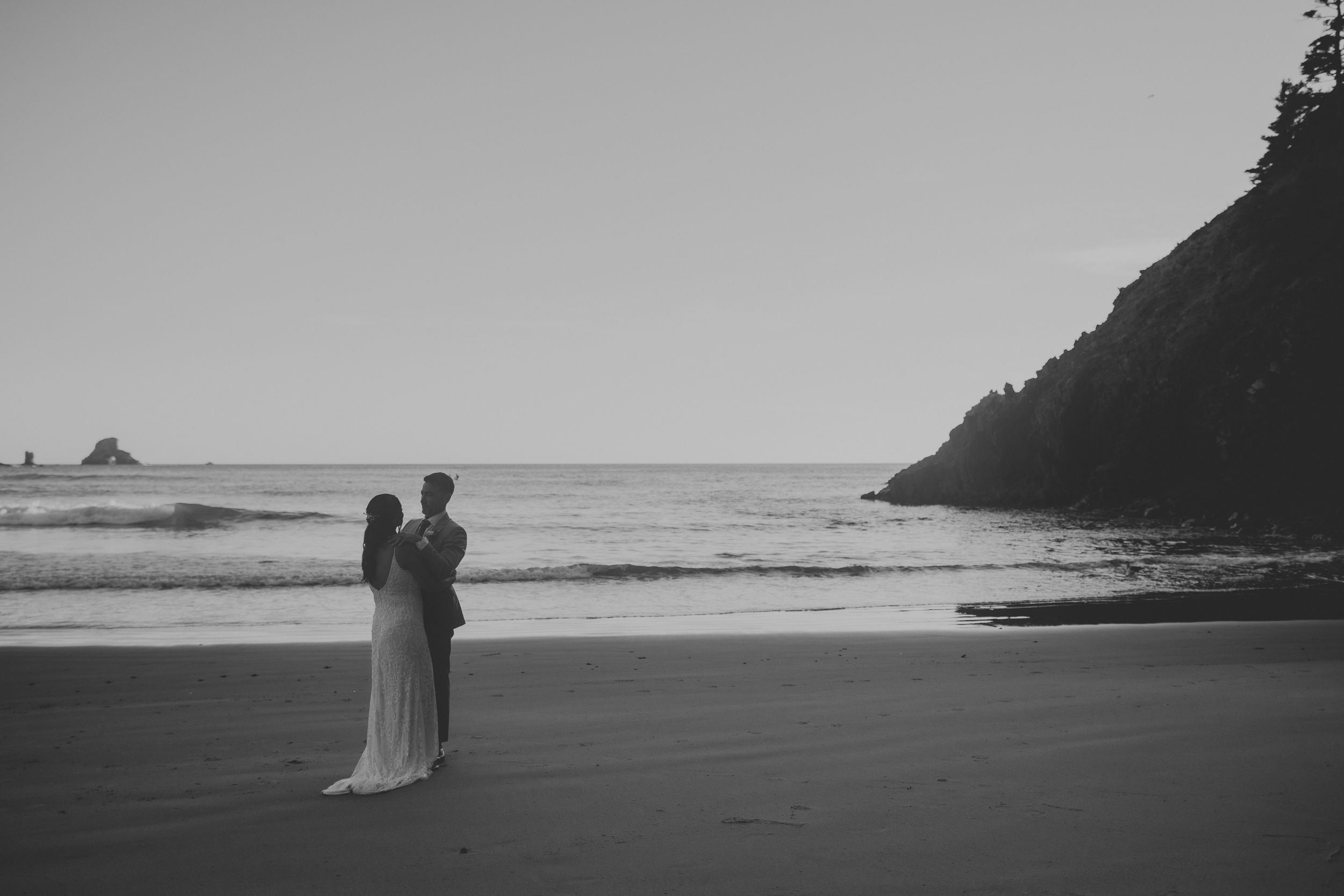 Black and white photo of a couple standing on a beach near the ocean with a rocky cliff in the background.