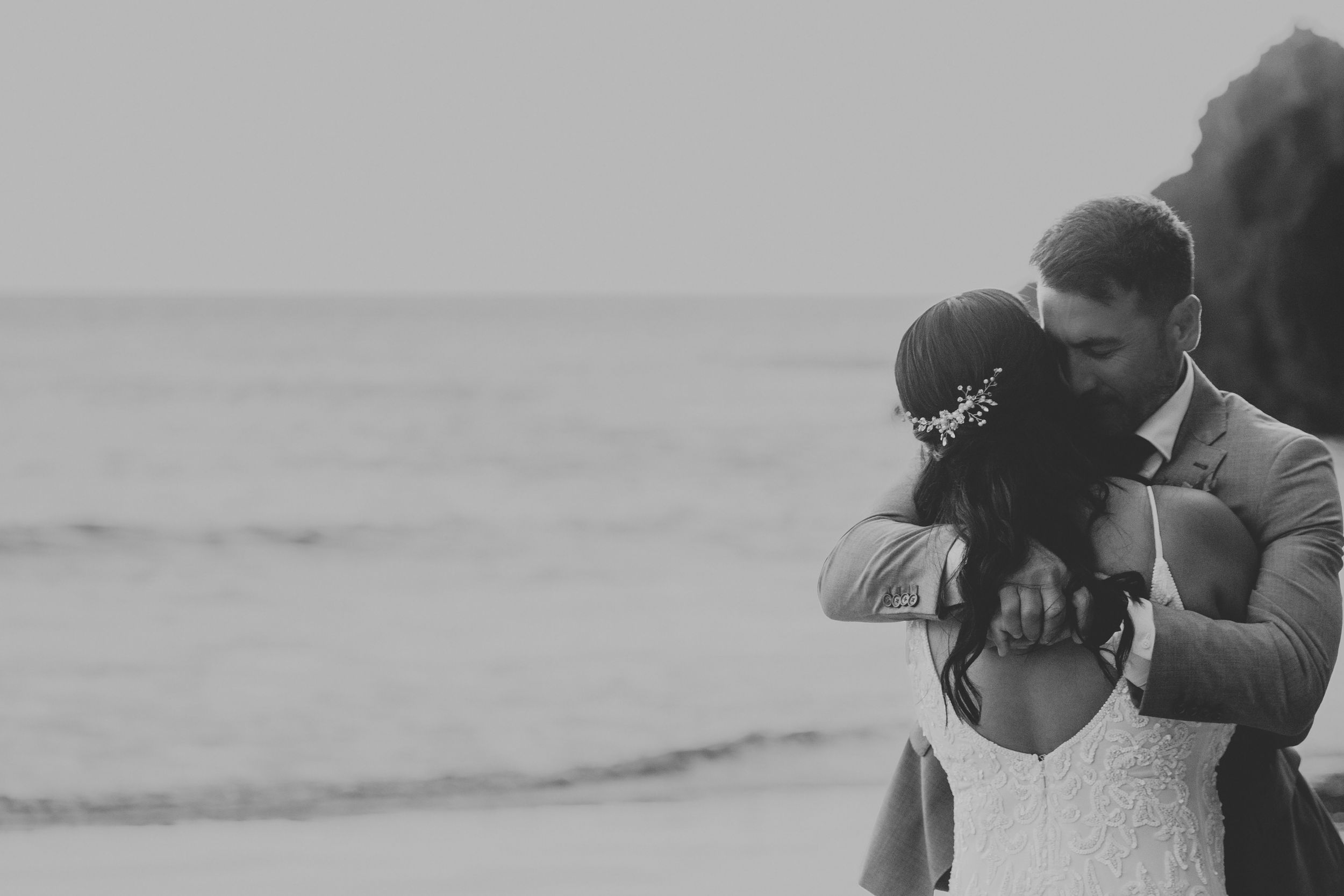 Black and white photo of a couple embracing on a beach in wedding attire, with ocean waves in the background.