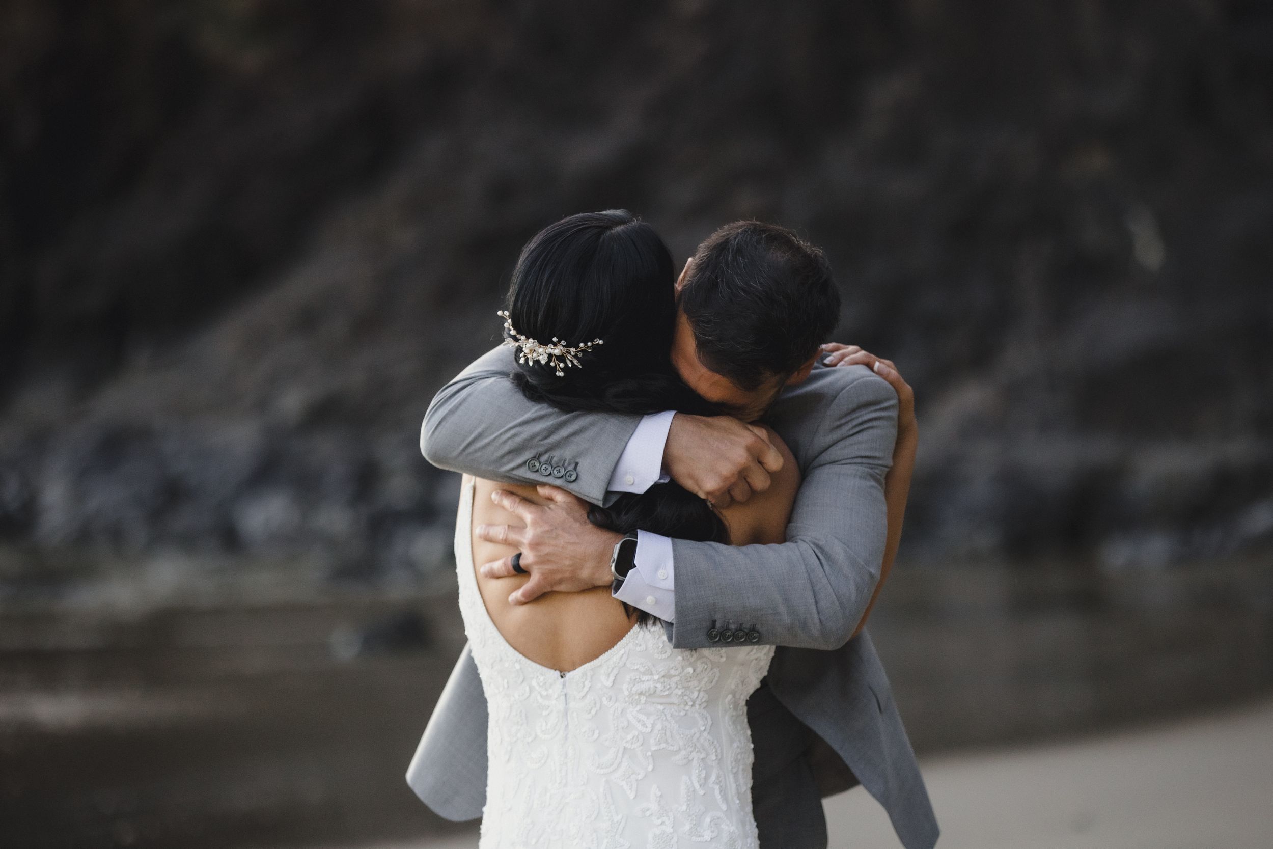 A bride and groom share a tender embrace on a beach, the bride in a lace gown and groom in a gray suit.