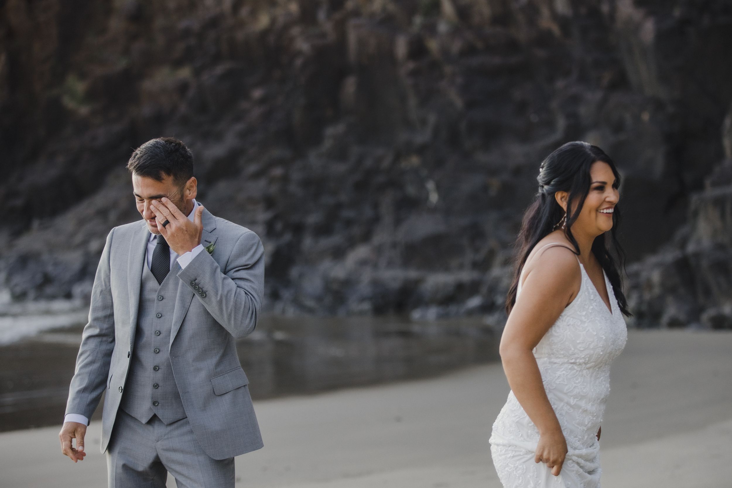Emotional groom wipes tears while bride smiles joyfully during a beach wedding first look moment.