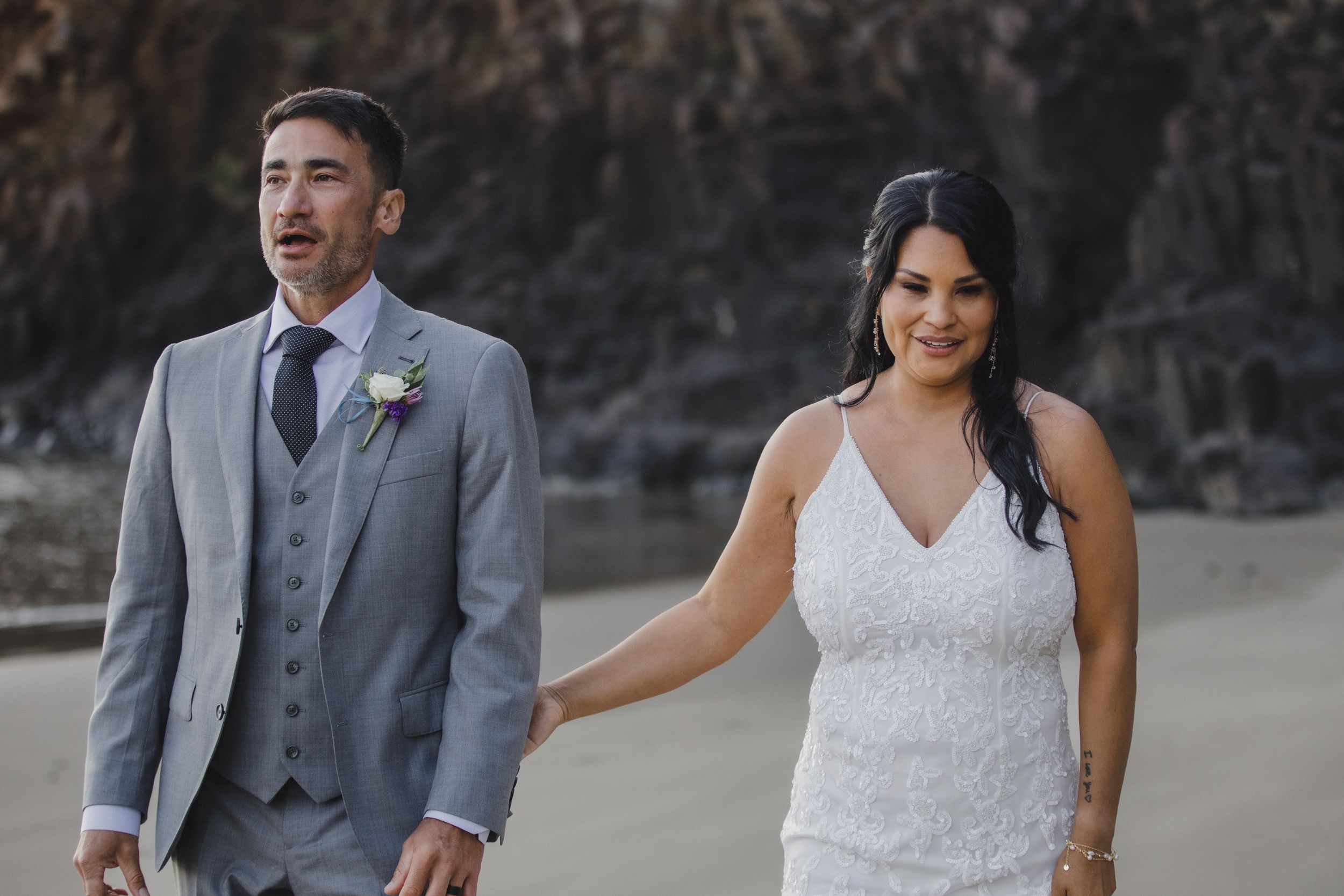Bride and groom walk hand in hand on a sandy beach, dressed in elegant wedding attire with cliffs behind them.