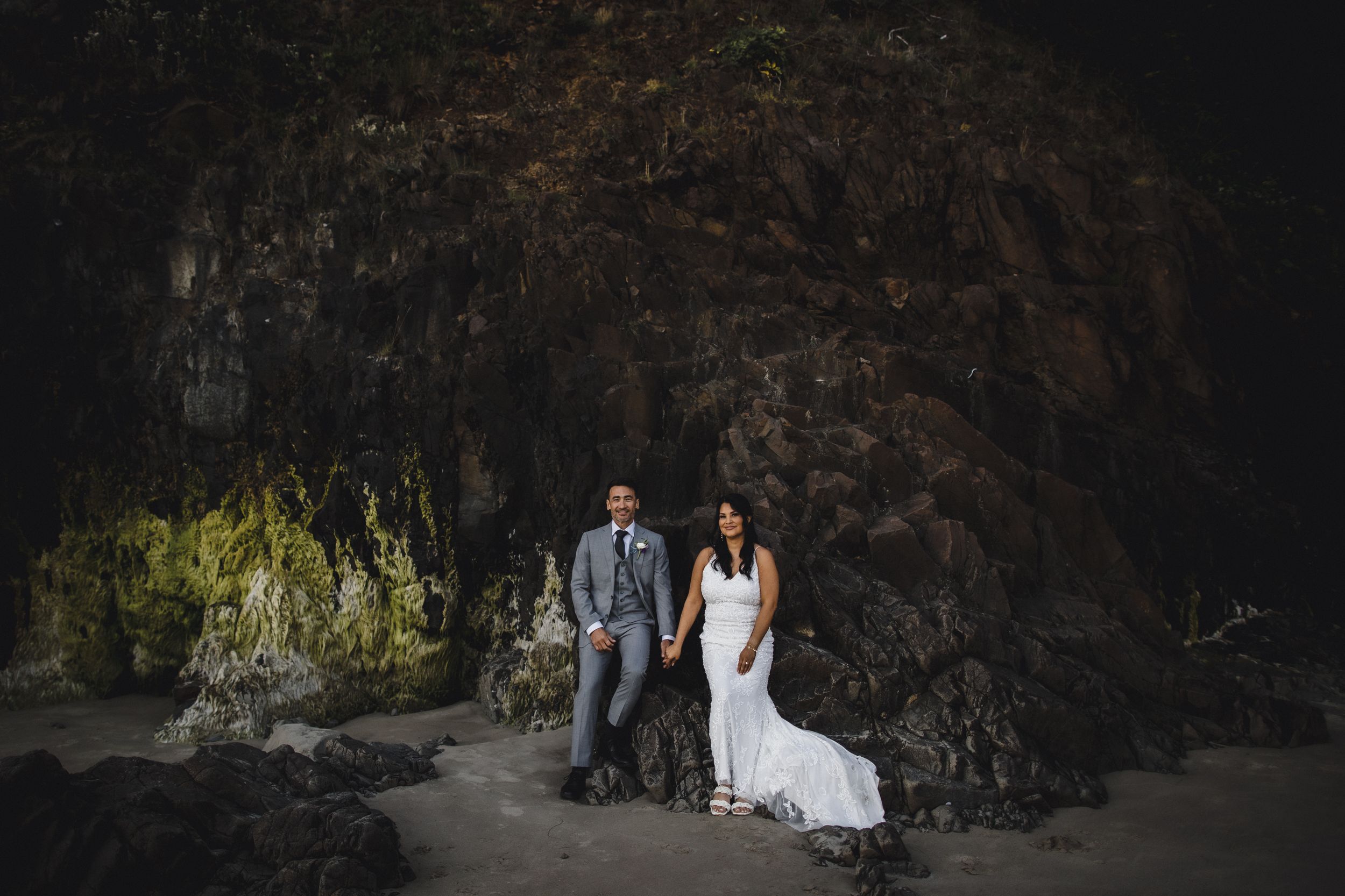 Bride and groom pose hand in hand on a dark sandy beach surrounded by towering mossy rock formations.