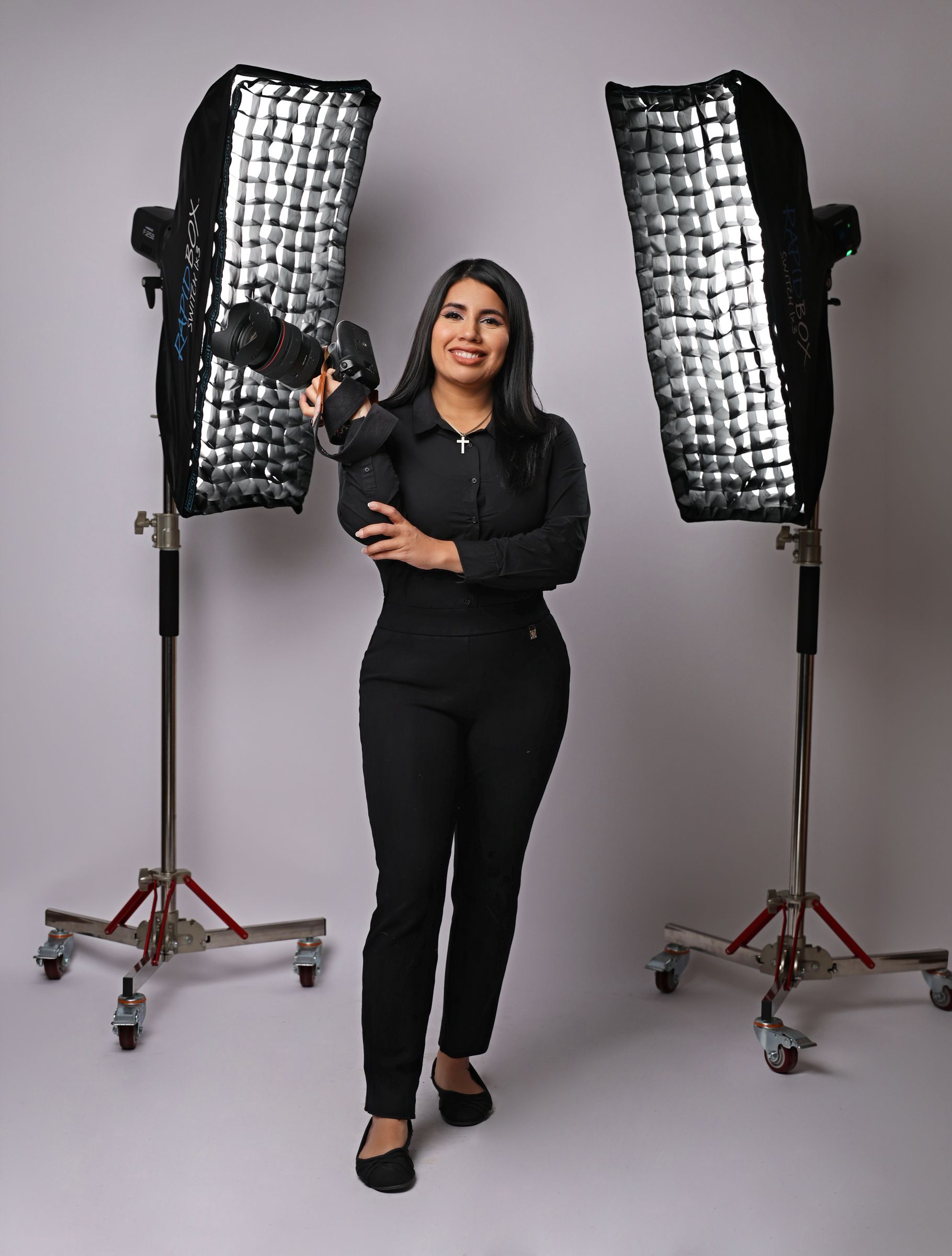 Female photographer in black outfit holding camera between two softbox studio lights on white background.