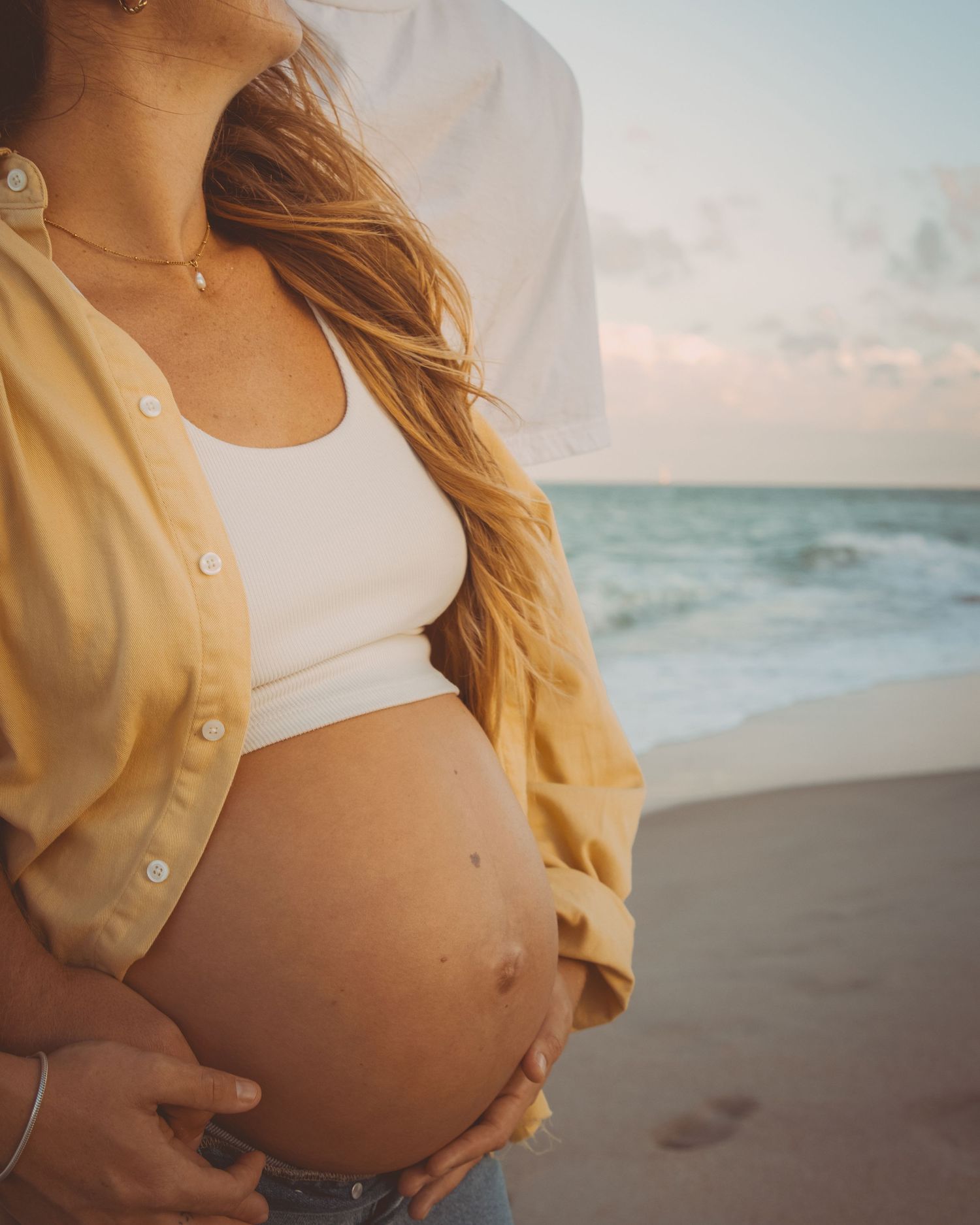 Pregnant woman in yellow shirt and white crop top cradling baby bump on a beach at sunset.
