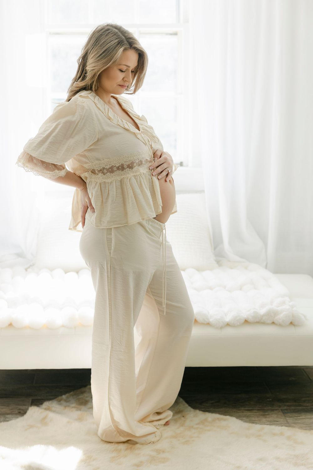 Pregnant woman in cream lace pyjama set posing sideways, cradling baby bump in bright white bedroom.