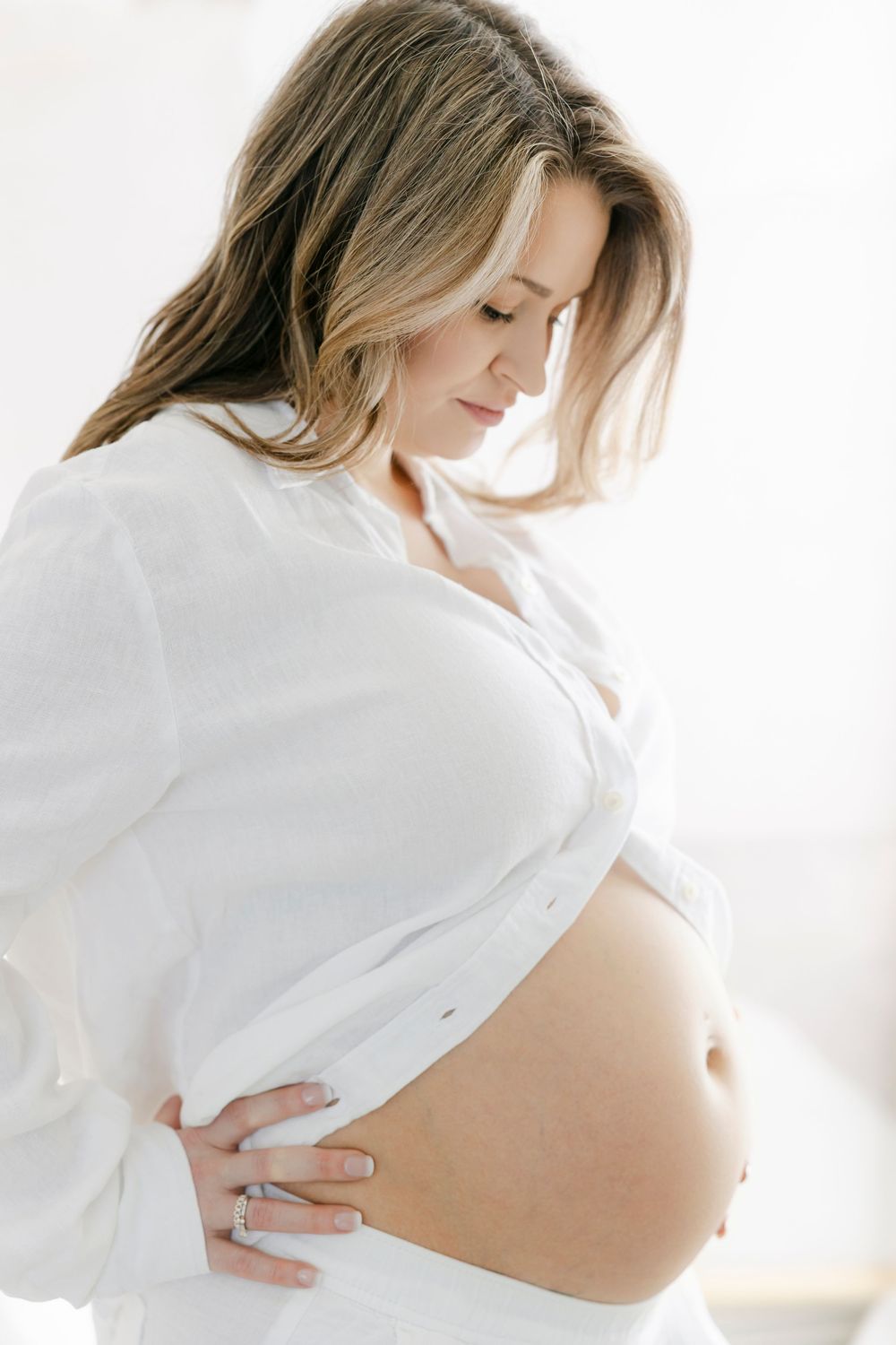 Pregnant woman in white shirt cradling her bare baby bump, looking down tenderly in a bright, airy setting.