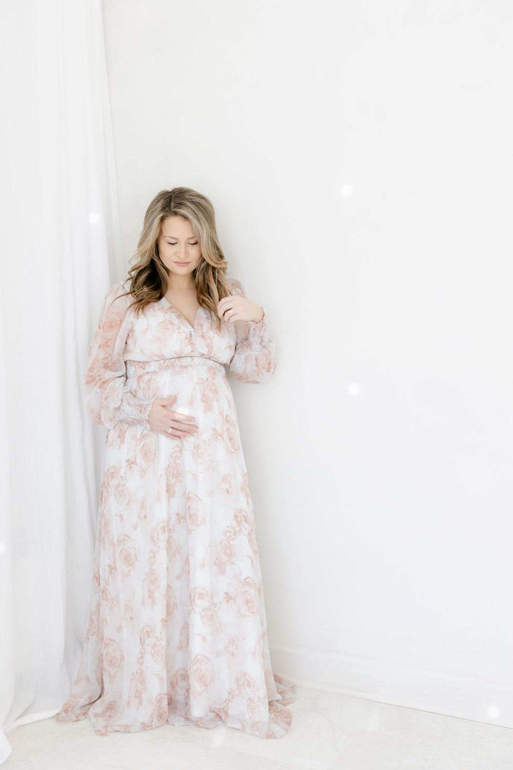 Pregnant woman in floral maxi dress posing gracefully in bright white studio for maternity photo session.
