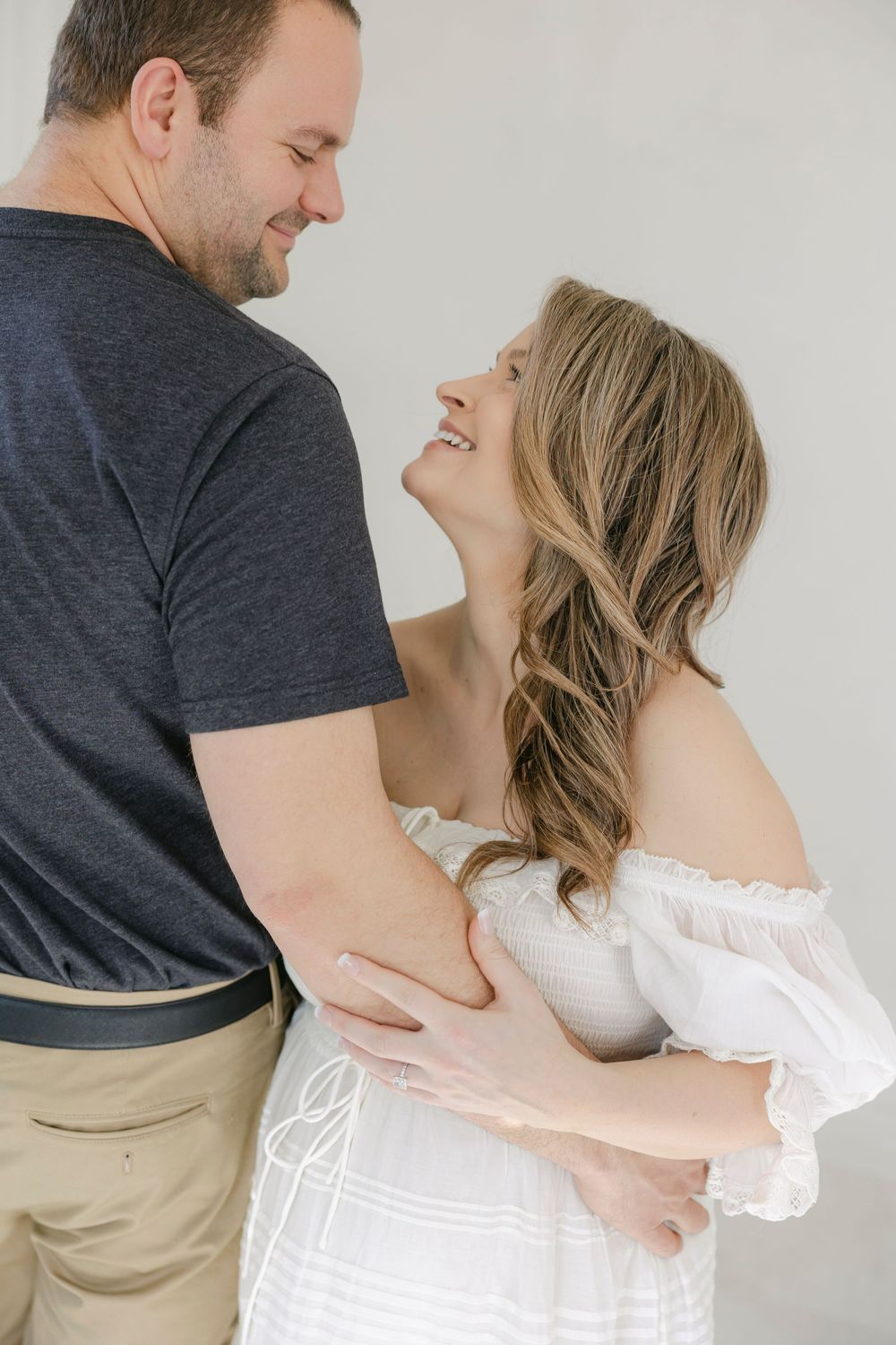 A smiling couple embraces tenderly, woman in white off-shoulder dress gazing up at man in navy shirt and khaki pants.