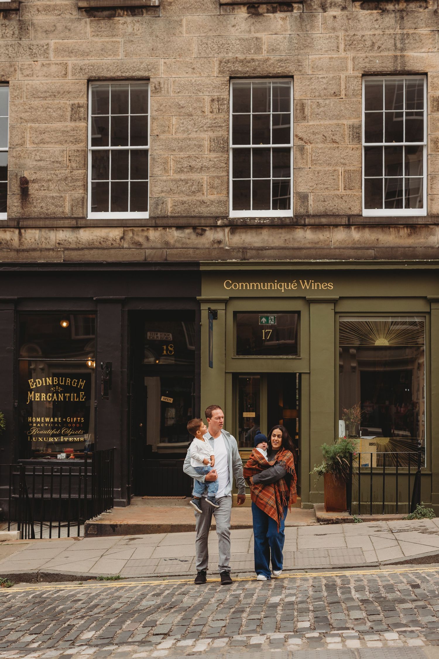 A family stands on cobblestone streets outside Edinburgh Mercantile and Communiqué Wines shops in Scotland.