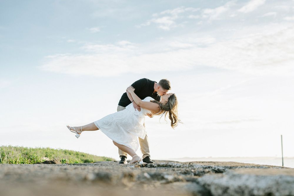 Couple sharing a romantic dip kiss outdoors on rocky ground with a blue sky backdrop.