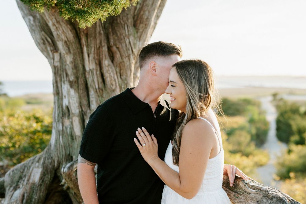 Couple sharing a romantic moment near a large tree outdoors, woman wearing white dress with engagement ring visible.