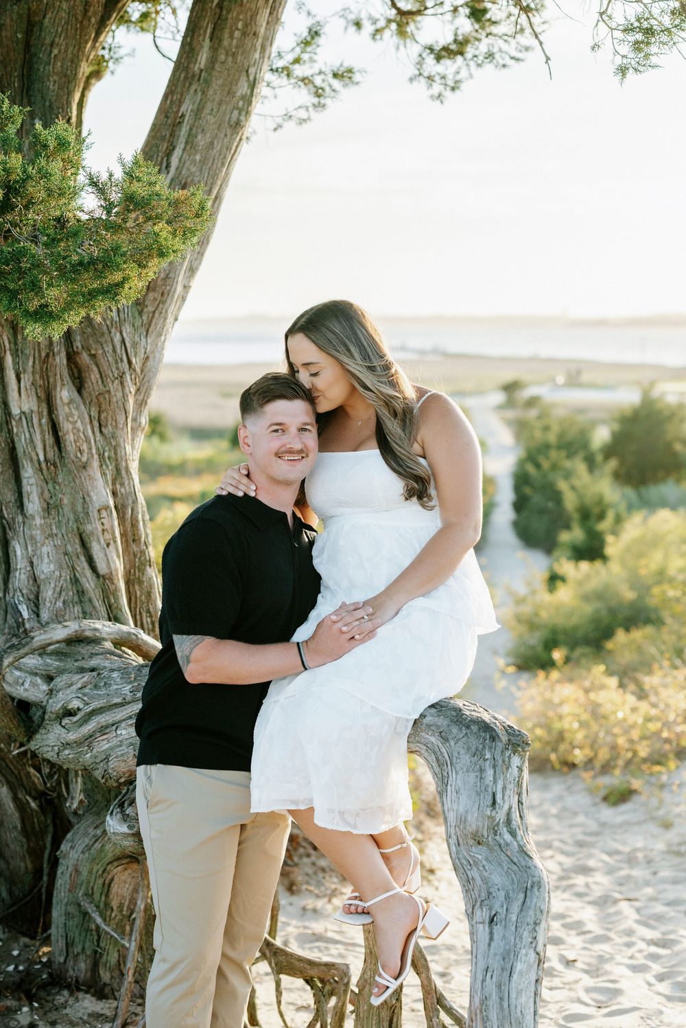 Smiling couple posing by a weathered tree on a sandy beach, woman in white dress at sunset.