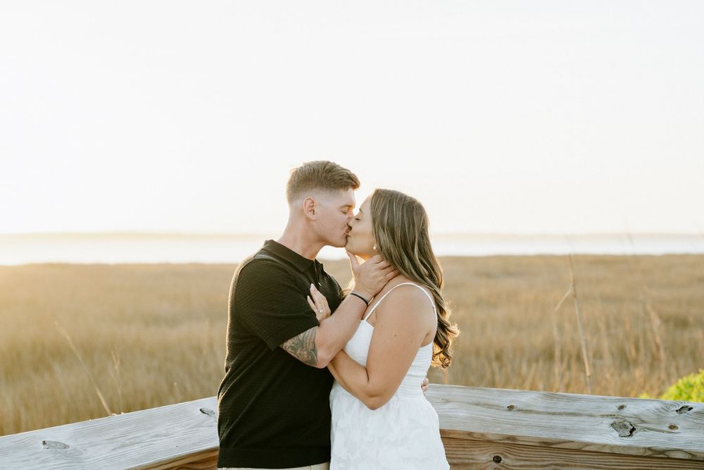 Couple sharing a romantic moment at golden hour in a coastal field, man in black shirt and woman in white dress.