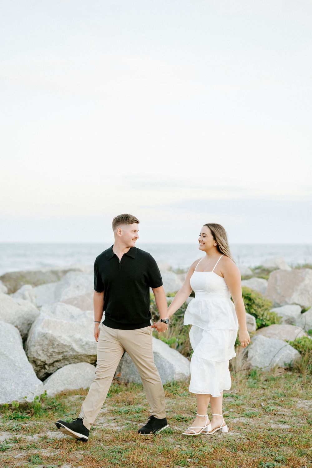 Couple holding hands and smiling at each other near rocky shoreline, man in black polo and woman in white dress.