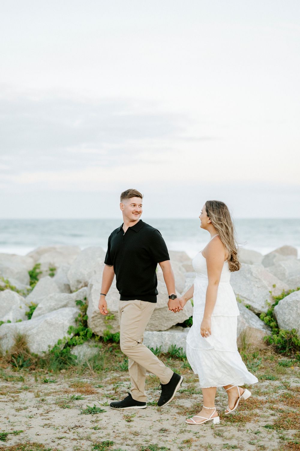 Couple holding hands walking on a rocky beach, man in black shirt and woman in white dress with ocean backdrop.