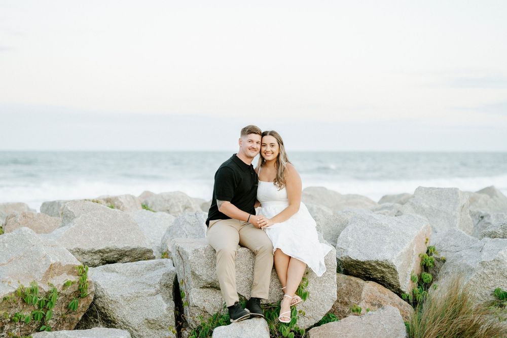 Happy couple sitting on large rocks at the beach, embracing and smiling with ocean waves behind them.
