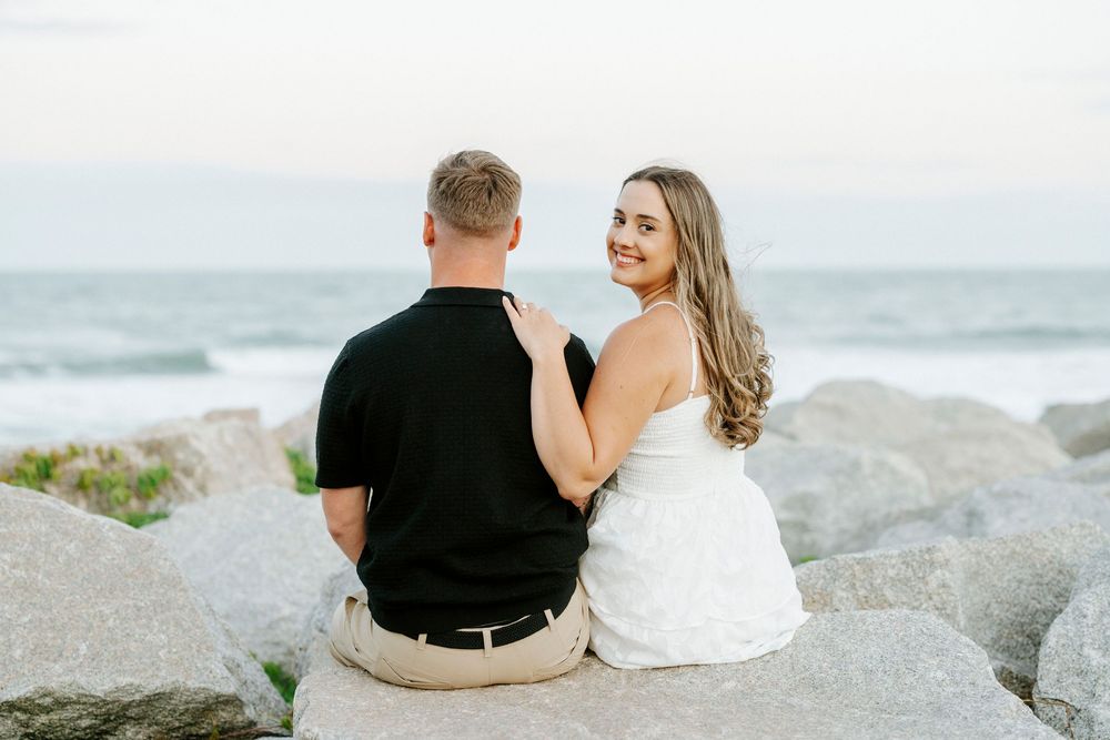 Couple sitting on rocks by the ocean, woman in white dress smiling back at camera, man in black shirt facing the sea.