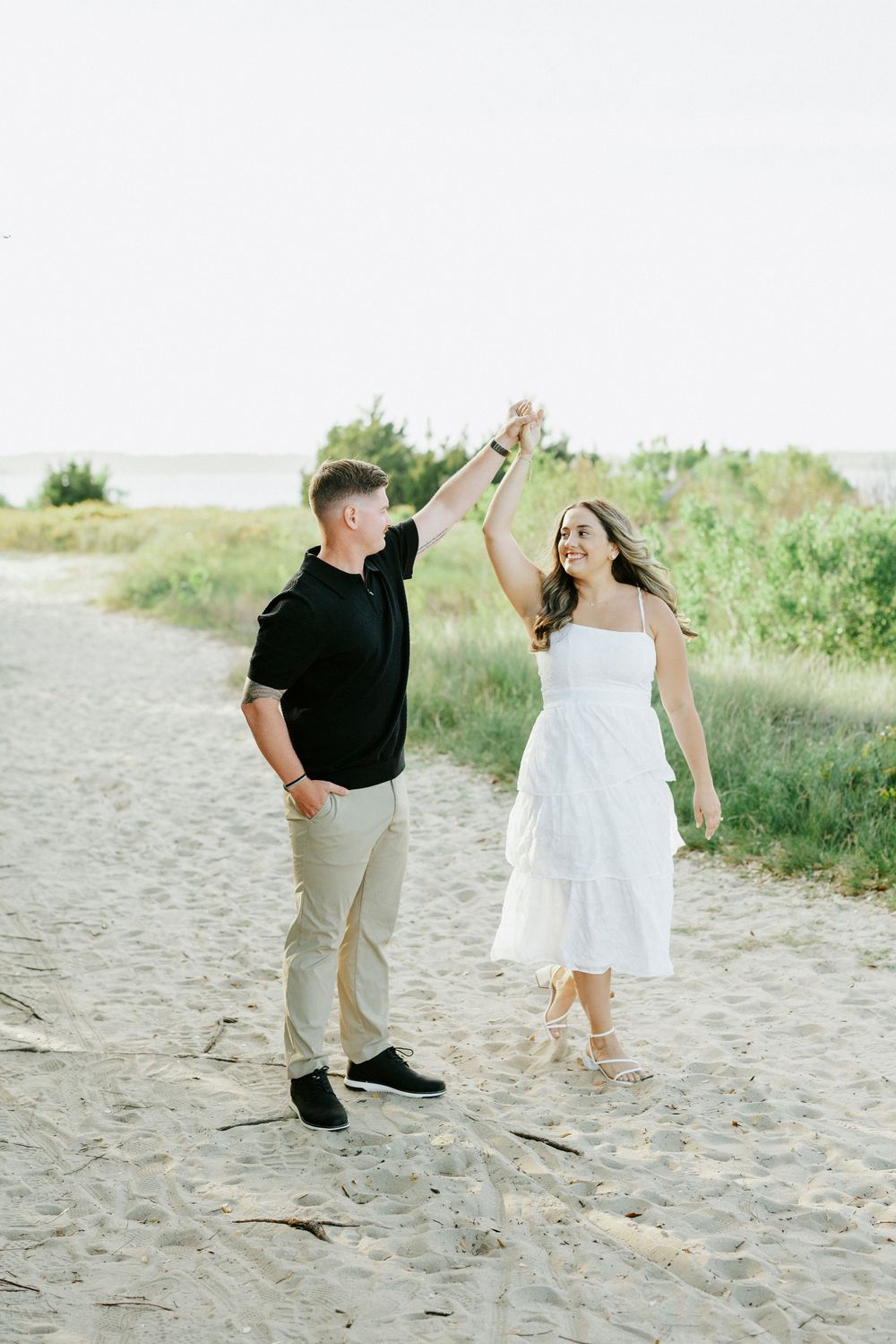 A couple dances on a sandy beach path, woman in white dress, man in black shirt, with green dunes behind them.
