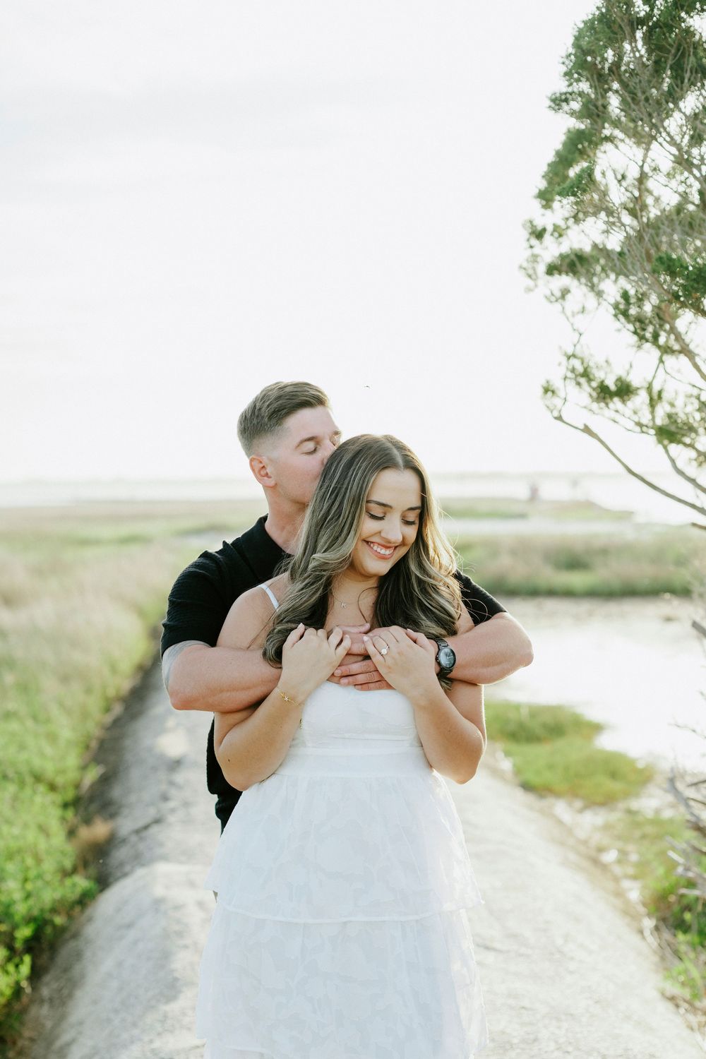 A couple embraces on a coastal path, man in black hugging woman in white dress, smiling outdoors.