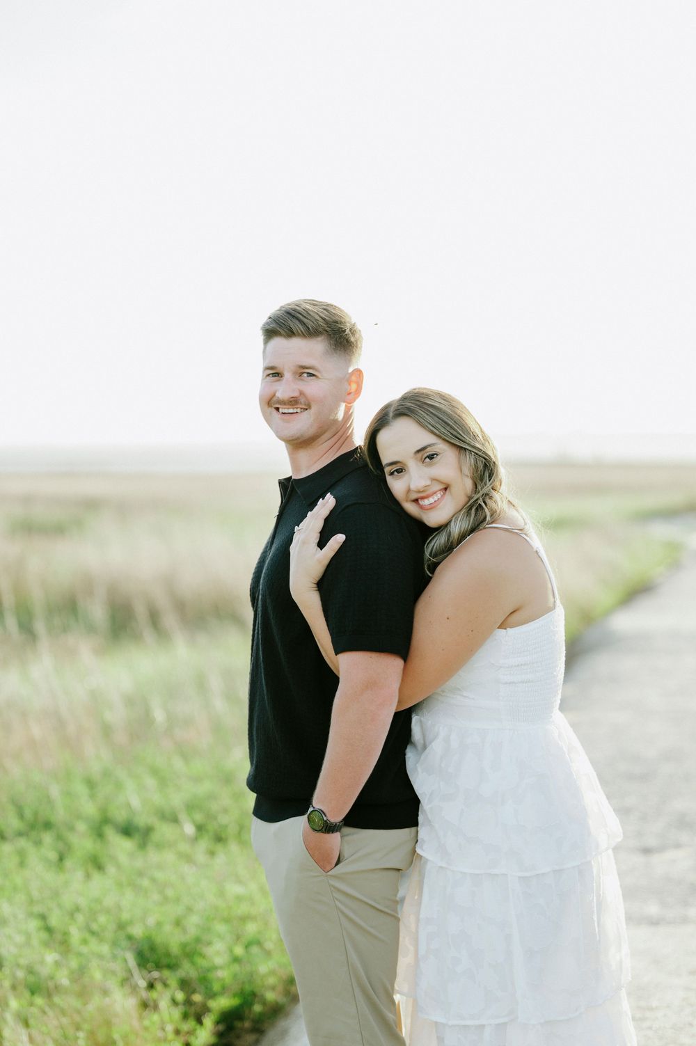 Couple posing outdoors, woman in white dress embracing man in black polo shirt with green field background.