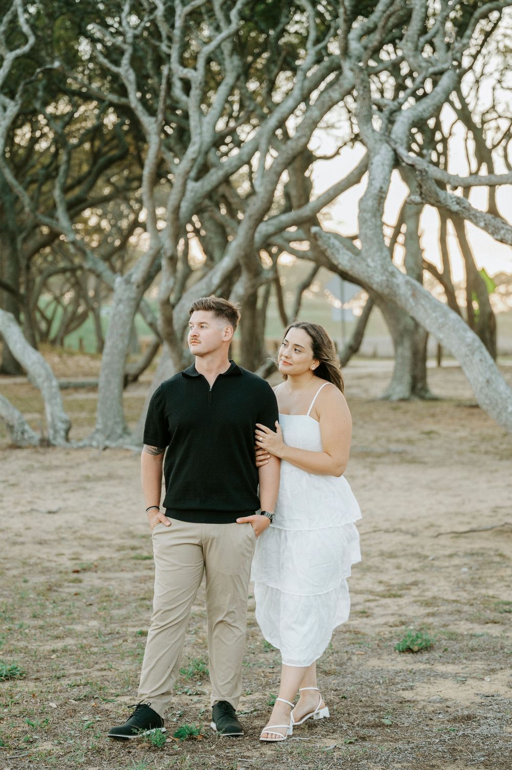Couple posing outdoors among twisted trees, man in black polo and woman in white dress, golden hour light.