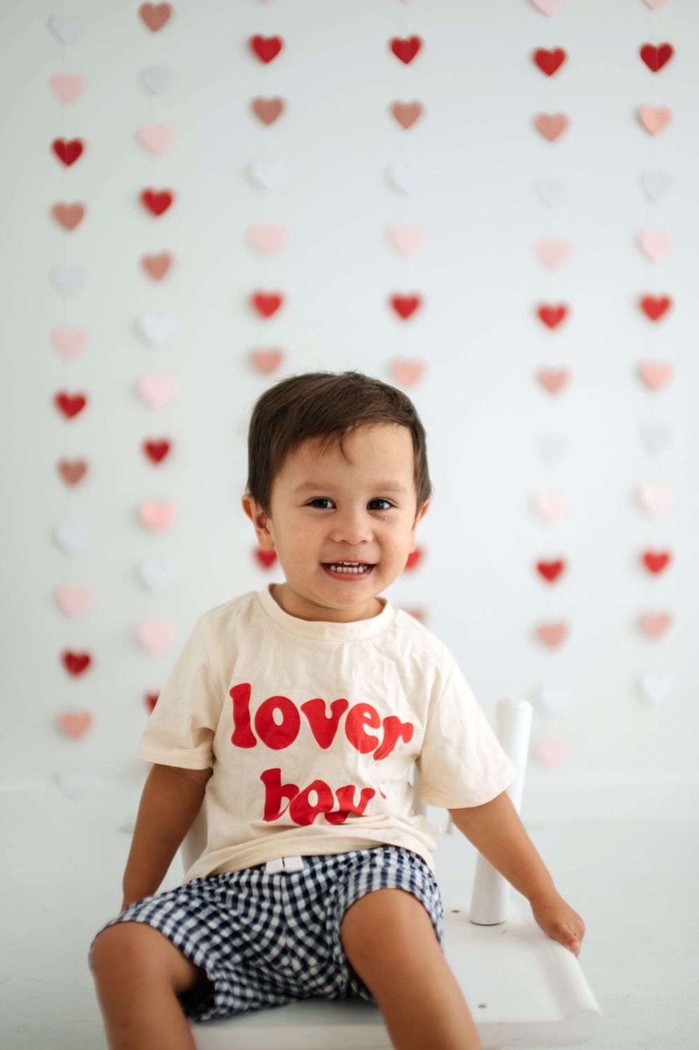 Smiling toddler boy wearing a cream “lover boy” t-shirt sits in front of a heart garland backdrop.
