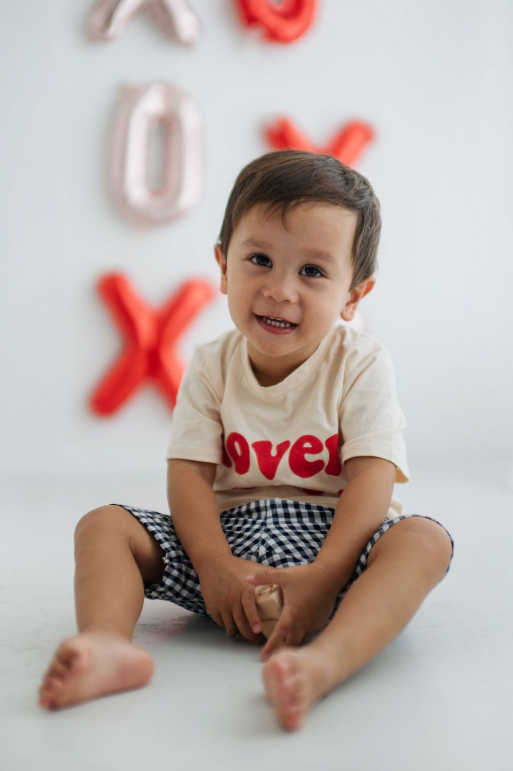 Smiling toddler boy sitting on floor wearing a love shirt and checkered shorts with XOXO balloons behind him.