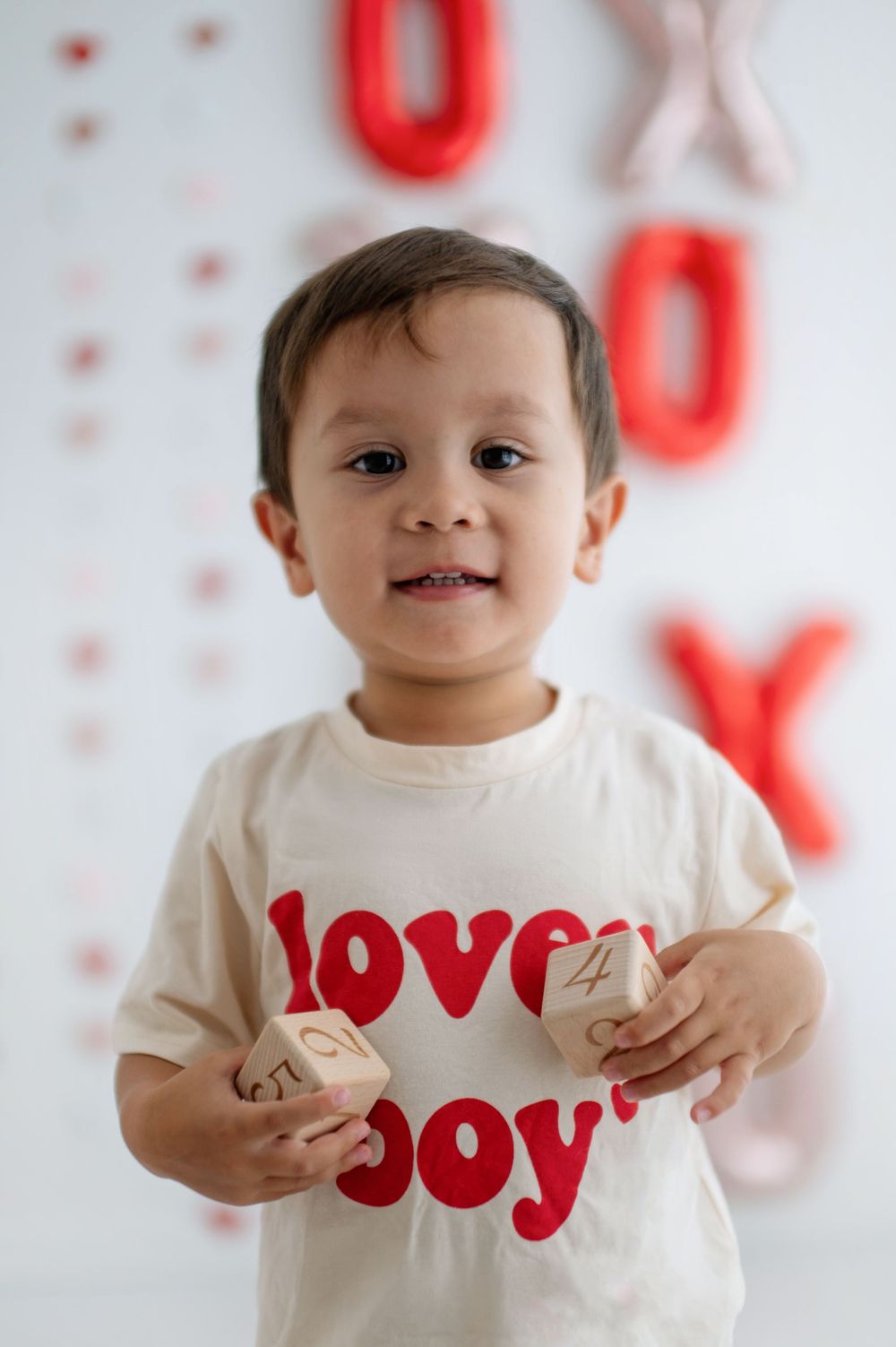 Toddler boy in a 'Lovely Boy' shirt holds wooden blocks, standing before red and pink XOXO balloon decorations.