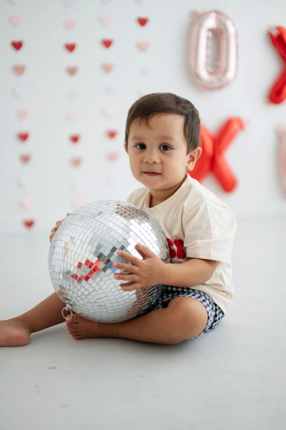 Toddler boy holding a disco ball, sitting on white floor with Valentine's Day heart decorations and XOXO balloons.