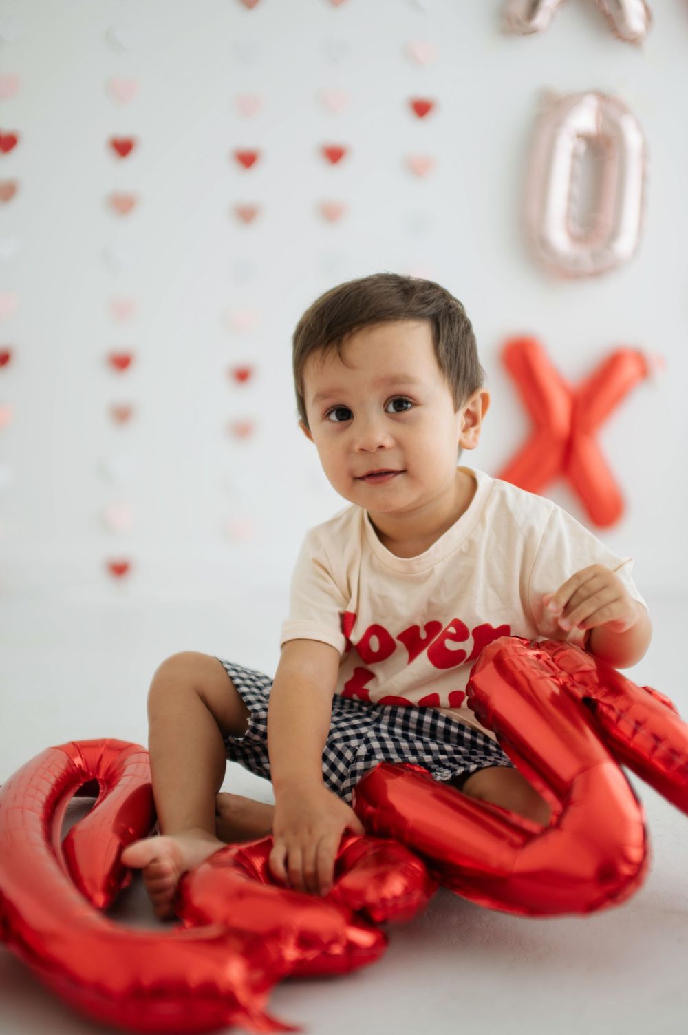 Toddler boy sitting with red heart balloons on Valentine's Day with XO decorations on white backdrop.