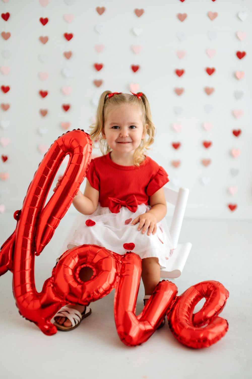 Smiling toddler girl in red and white dress with red LOVE balloons and heart decorations for Valentine's Day.