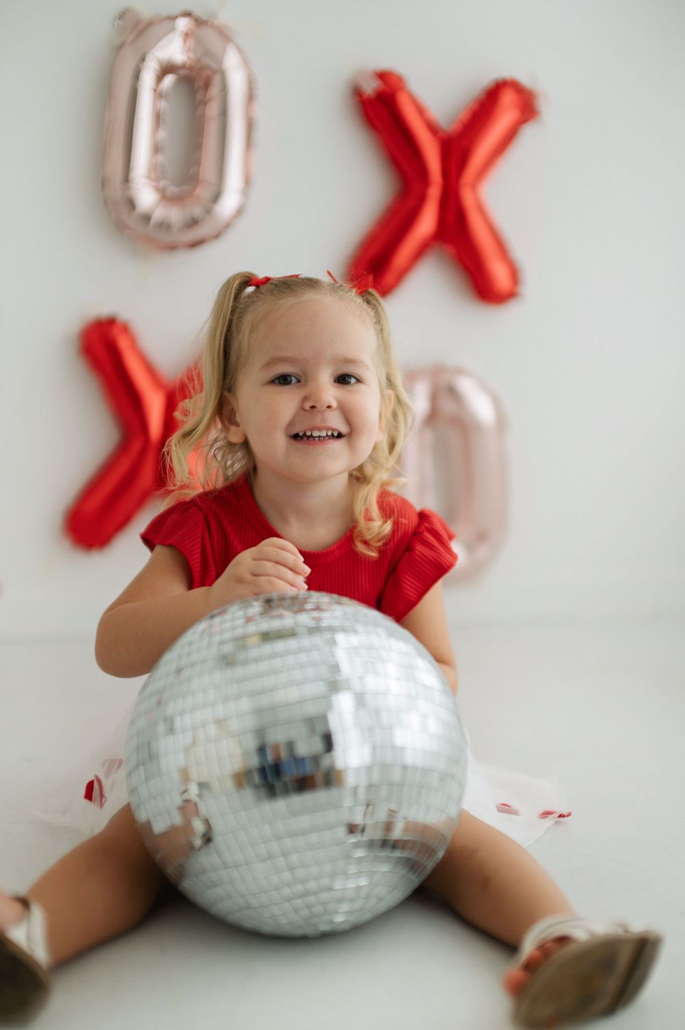 Smiling toddler girl in red dress holding disco ball with XOXO balloons on white background.
