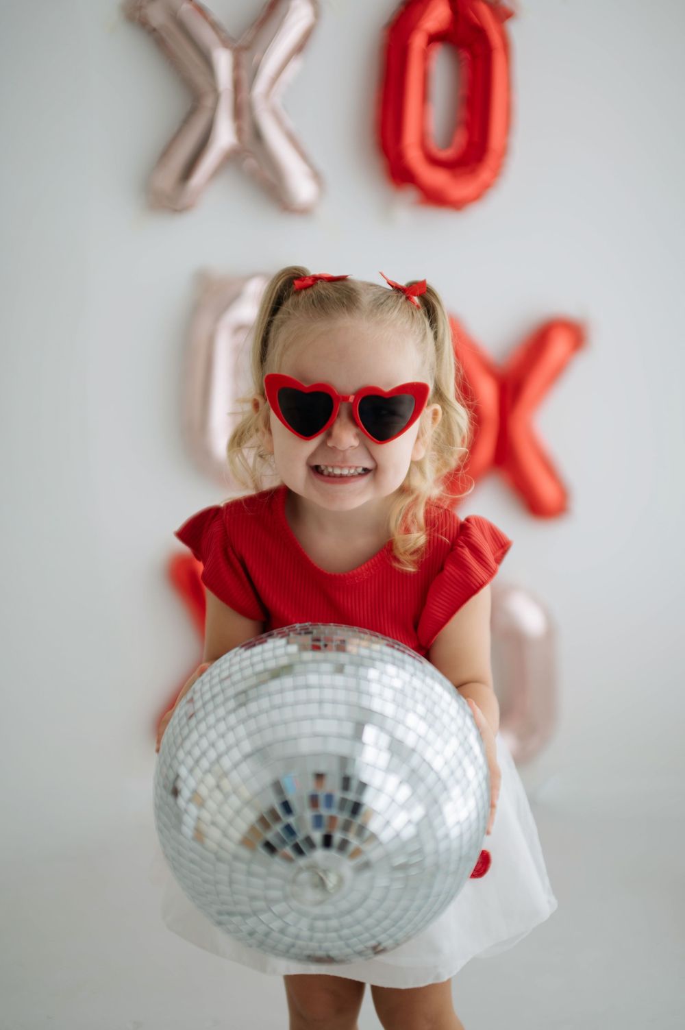 Young girl in red heart sunglasses holds disco ball, wearing red dress, with XOXO balloons on white wall.