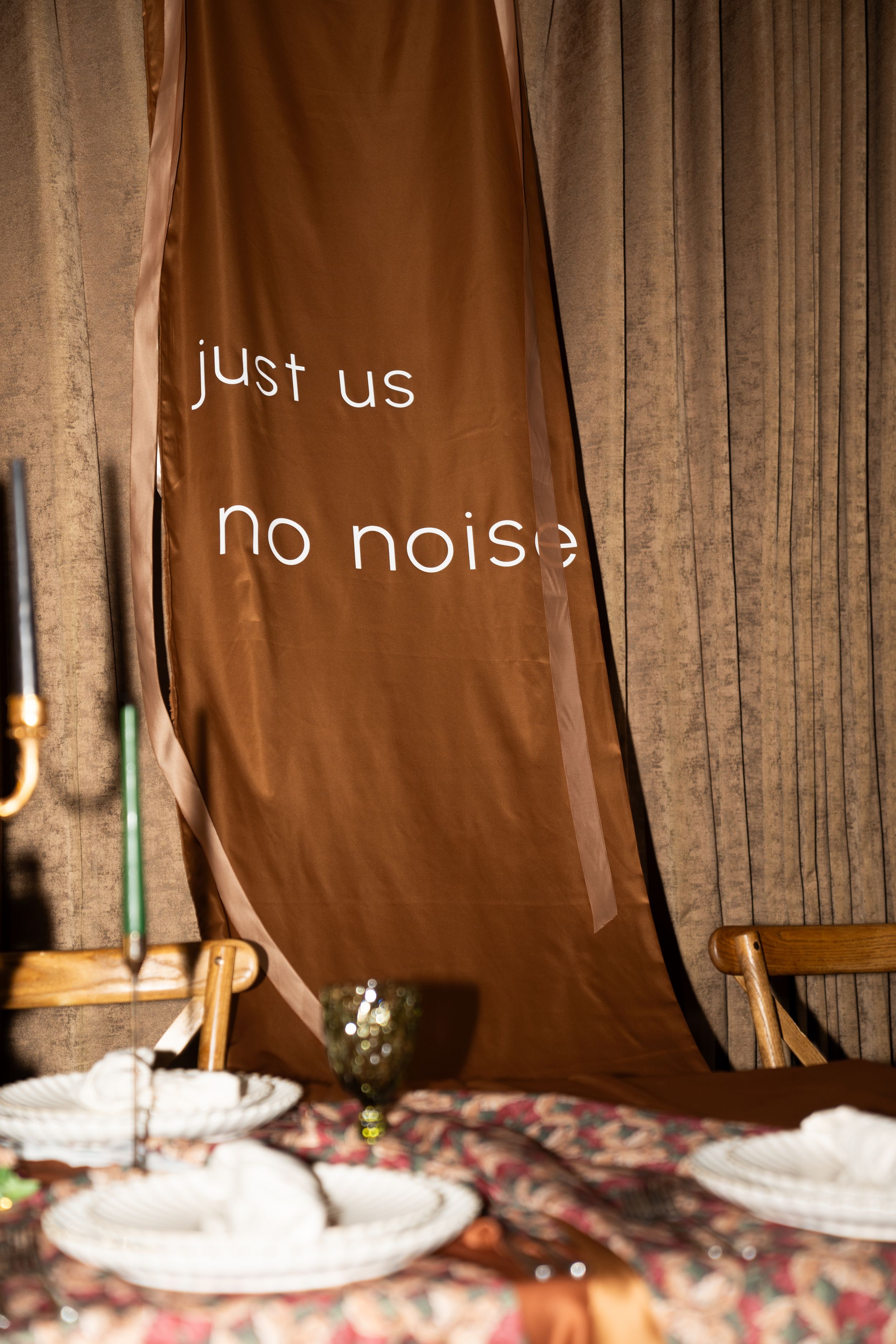 Brown fabric banner reading 'just us no noise' hanging above a decorated dining table with plates and candles.