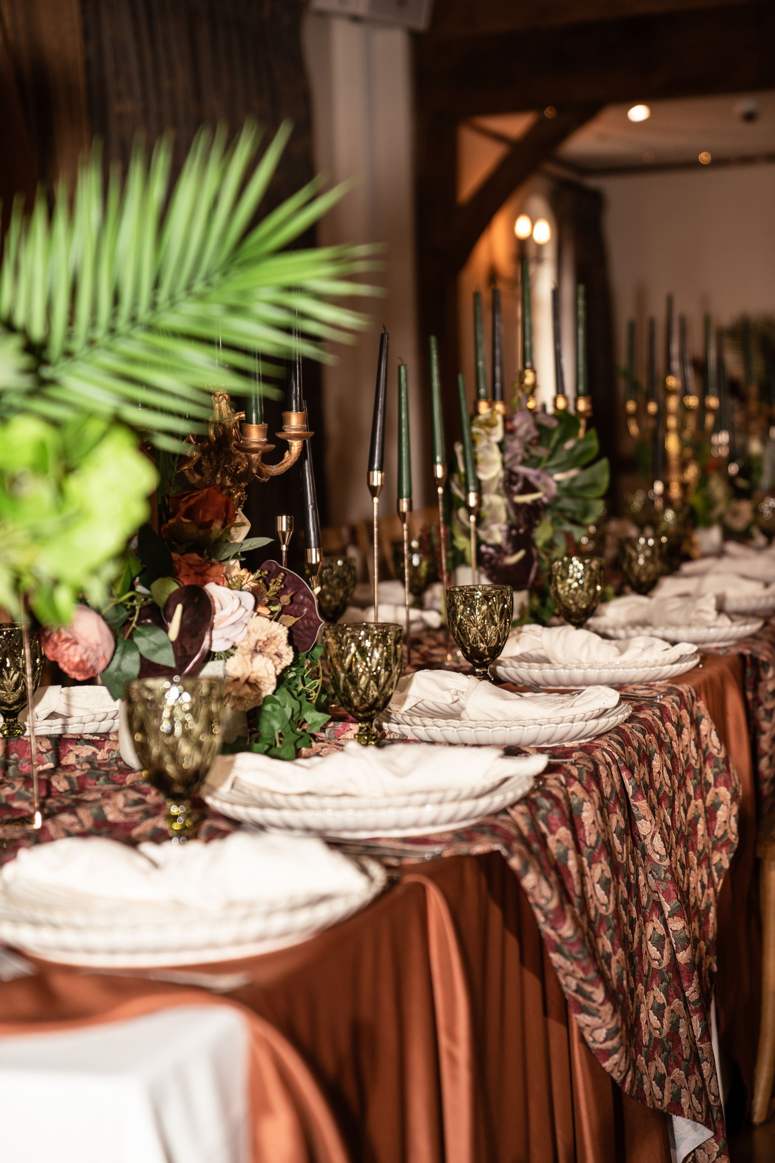 Elegant wedding reception table with dark candles, tropical greenery, floral arrangements, and ornate place settings on rich brown satin.
