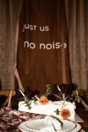 White wedding cake with orange flowers and dark leaves, displayed before a brown backdrop reading just us no noise.