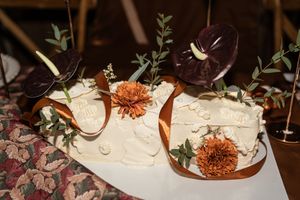 Two white wedding cakes decorated with rust ribbons, burgundy flowers, and eucalyptus greenery on a floral tablecloth.