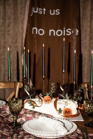 Elegant wedding table with black and green taper candles, white cake, floral runner, and "just us no noise" backdrop.