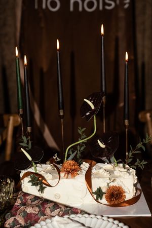 White wedding cake with orange chrysanthemums, dark anthurium flowers, and tall black candles on a decorated table.