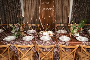 Elegant wedding reception table with floral tablecloth, gold candlesticks, white plates, and rustic wooden chairs.